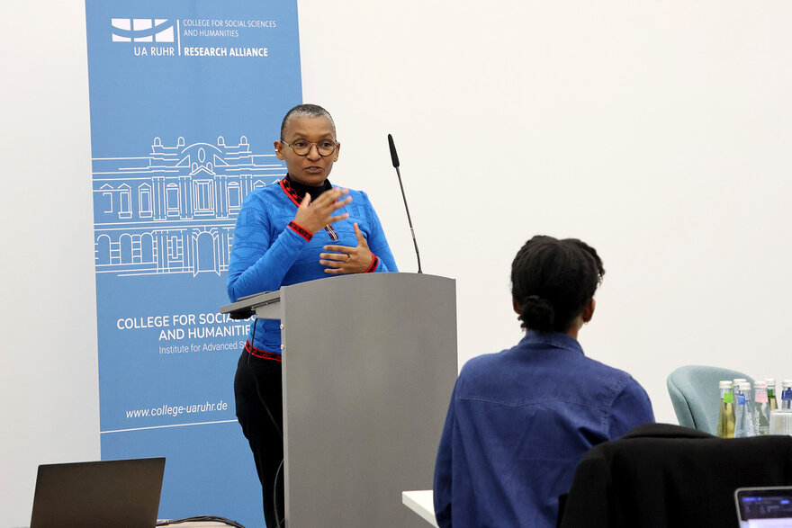 Matebeni giving a lecture, standing in front of a speaker's desk, behind her the blue College banner